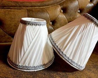 Könnte beinhalten: Zwei weiße Lampenschirme mit einem plissierten Design und einem silbernen Besatz. Die Lampenschirme liegen auf einer braunen Lederoberfläche.