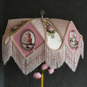 Pu&ograve; includere: Un paralume rosa e bianco con una frangia di perline e disegni floreali. Il paralume presenta una torta rosa e una tazza da t&egrave; con rose.