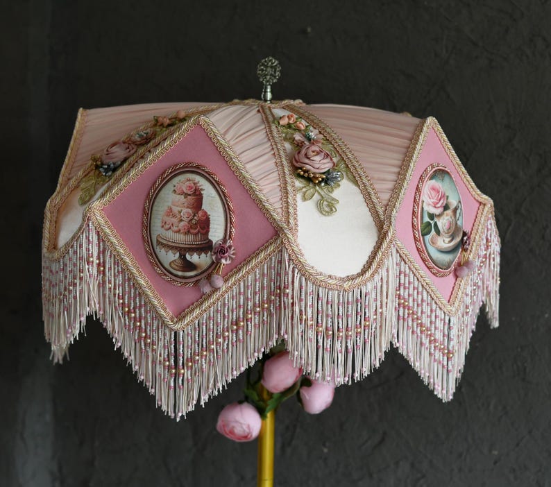 Pu&ograve; includere: Un paralume rosa e bianco con una frangia di perline e disegni floreali. Il paralume presenta una torta rosa e una tazza da t&egrave; con rose.