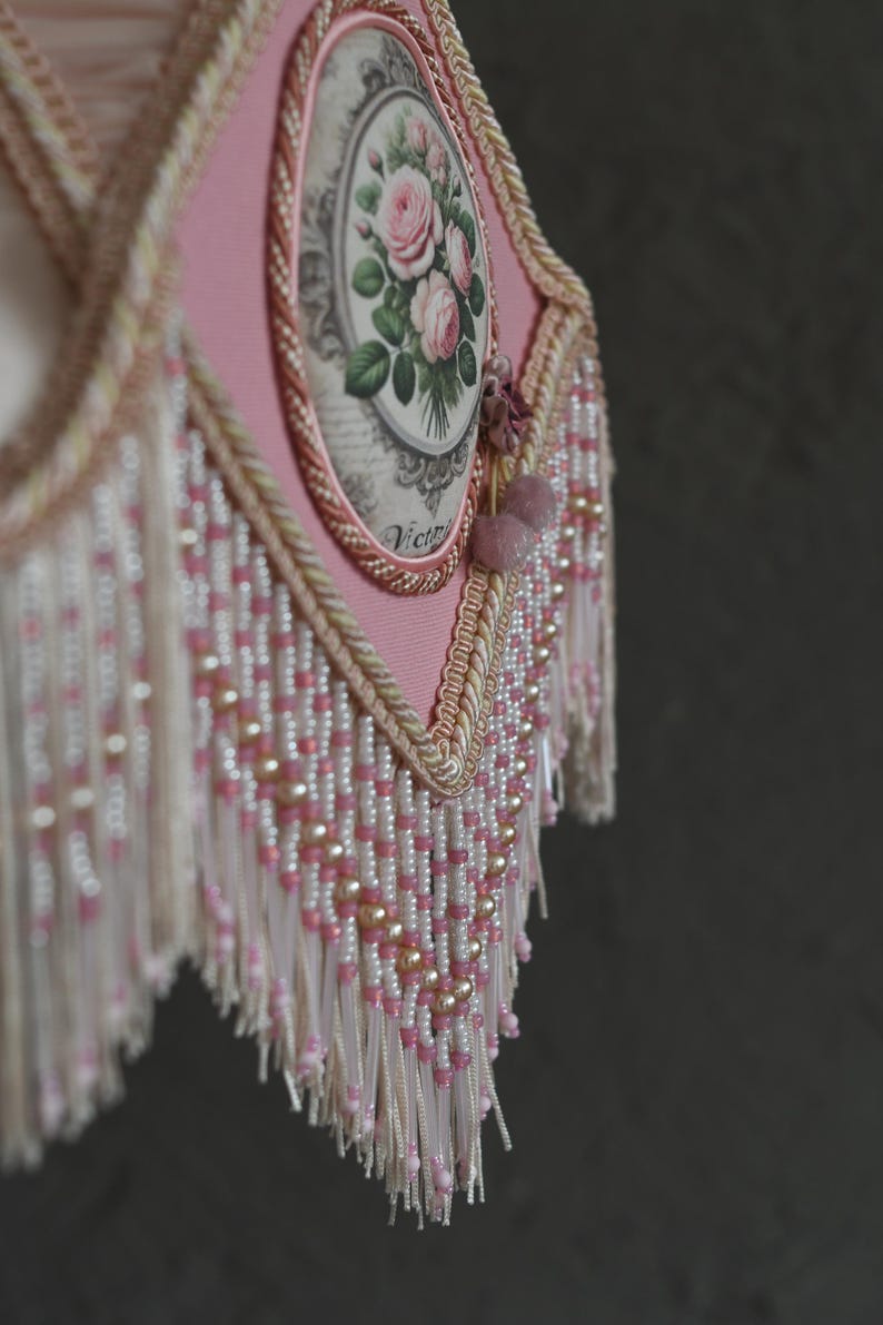 Pu&ograve; includere: Un paralume rosa con un motivo floreale e una frangia di perline. Il motivo floreale presenta rose e la parola "Victoria" &egrave; stampata sul paralume.