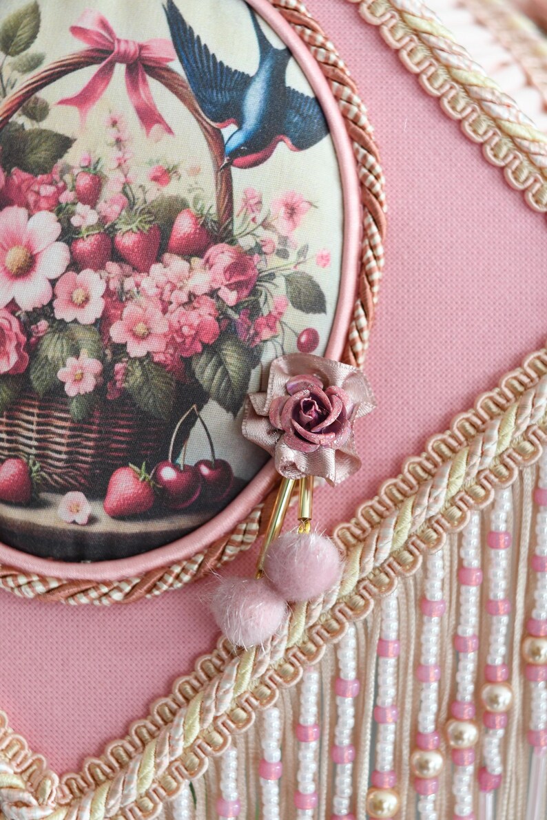 Pu&ograve; includere: Un paralume rosa con un pannello rotondo in tessuto con motivo floreale che presenta un uccello, ciliegie e fragole. Il pannello &egrave; circondato da una treccia marrone e una frangia bianca e dorata.