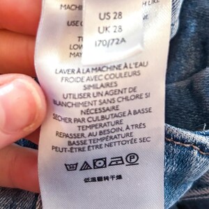 May include: A white clothing tag with care instructions and sizing information. The tag includes the sizes US 28 and UK 28. Washing instructions are in French. The tag is attached to a blue denim garment.