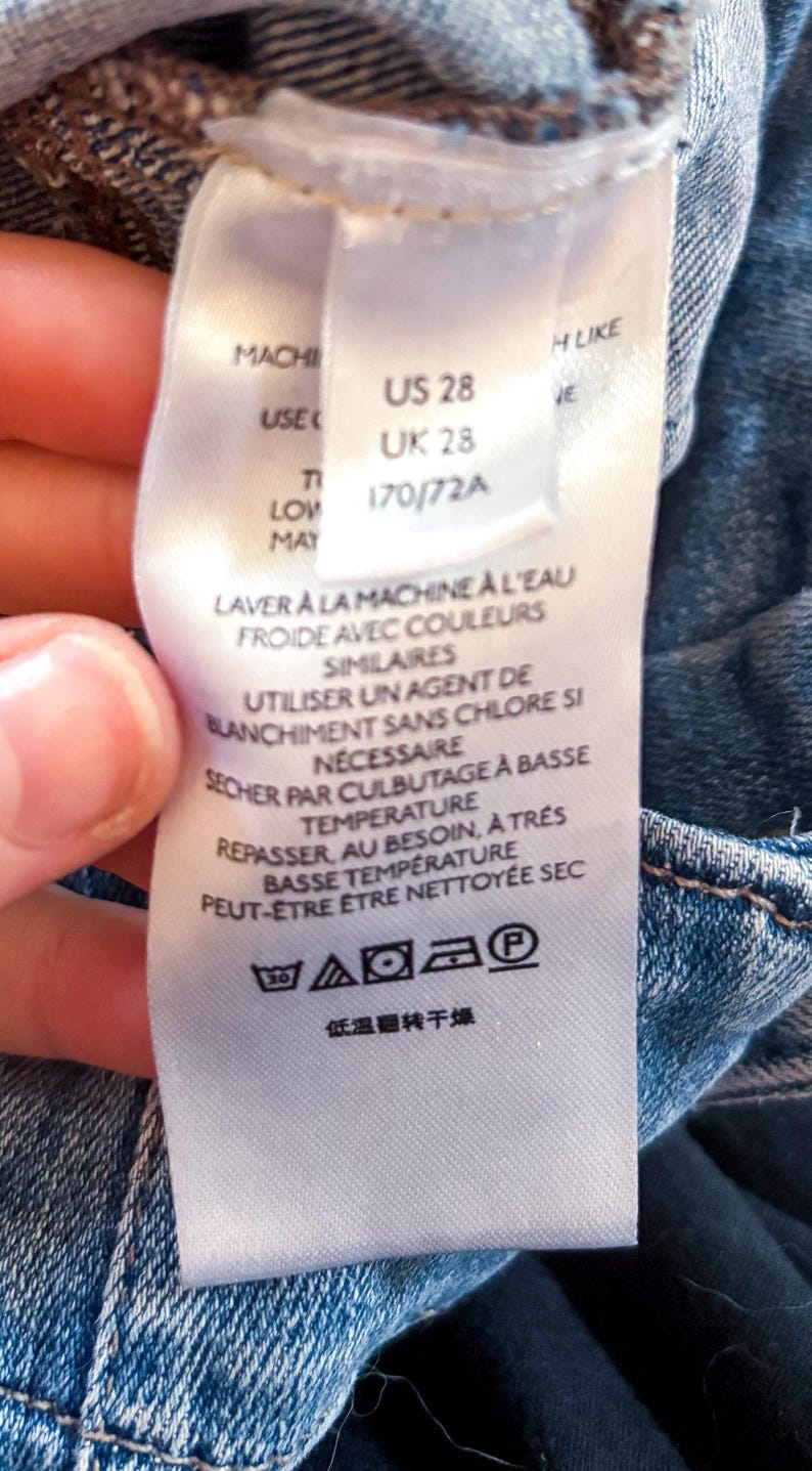 May include: A white clothing tag with care instructions and sizing information. The tag includes the sizes US 28 and UK 28. Washing instructions are in French. The tag is attached to a blue denim garment.