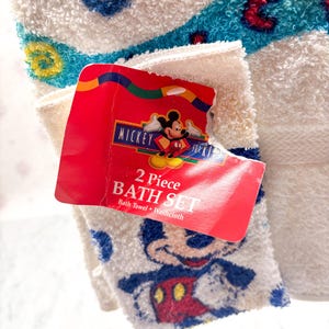 May include: A white 2-piece bath set featuring a bath towel and washcloth. The washcloth has a Mickey Mouse design. The set includes a red tag with the words "Mickey for Kids" and "2 Piece Bath Set". The towel has blue and red accents.