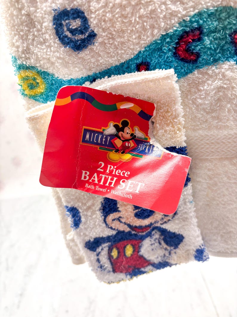 May include: A white 2-piece bath set featuring a bath towel and washcloth. The washcloth has a Mickey Mouse design. The set includes a red tag with the words "Mickey for Kids" and "2 Piece Bath Set". The towel has blue and red accents.