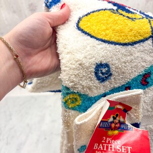 May include: A two-piece bath set featuring a white bath towel and washcloth with a Mickey Mouse design. The towel has a yellow, blue, and red color scheme. The set includes a tag that reads "Mickey for Kids".