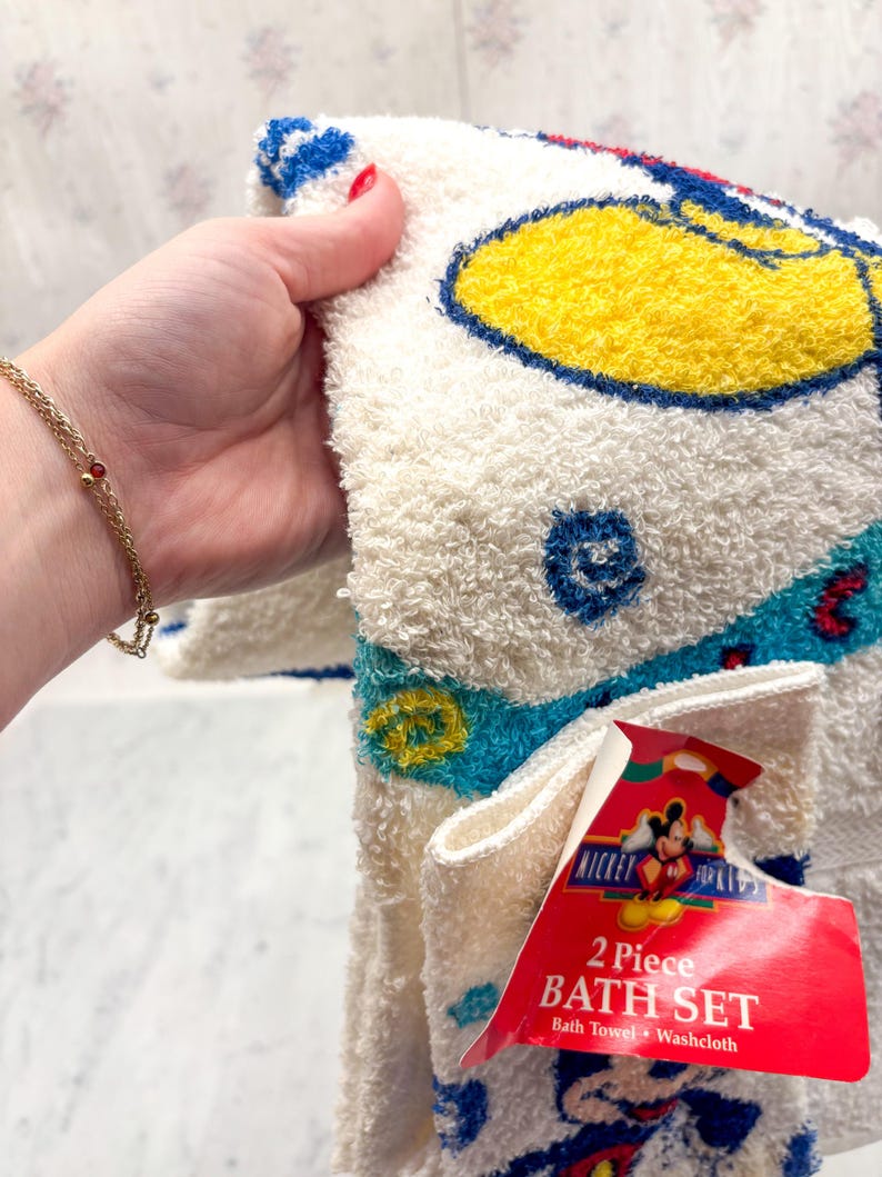 May include: A two-piece bath set featuring a white bath towel and washcloth with a Mickey Mouse design. The towel has a yellow, blue, and red color scheme. The set includes a tag that reads "Mickey for Kids".