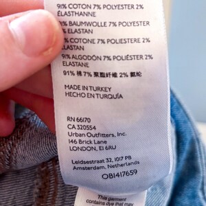 May include: A white clothing tag with text detailing fabric composition: 91% cotton, 7% polyester, and 2% elastane. The tag also includes the manufacturer's information, including the address in London, and a warning about potential dye transfer.
