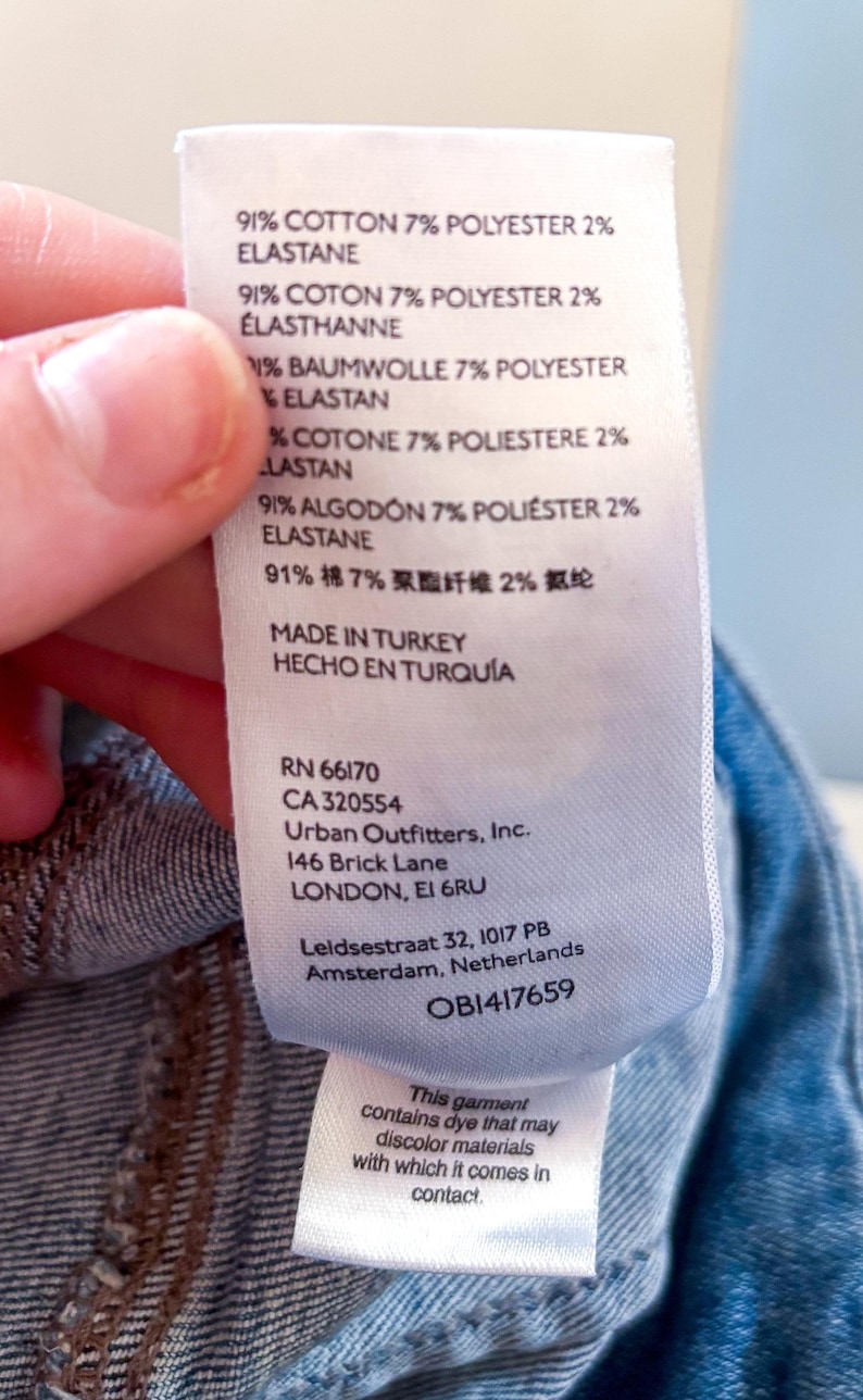 May include: A white clothing tag with text detailing fabric composition: 91% cotton, 7% polyester, and 2% elastane. The tag also includes the manufacturer's information, including the address in London, and a warning about potential dye transfer.