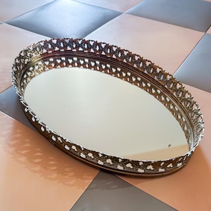 Puede incluir: Una bandeja de tocador ovalada con una base de espejo reflectante y un borde metálico decorativo con un intrincado patrón de filigrana calada. El marco de metal tiene un acabado dorado envejecido que rodea la superficie de vidrio central.