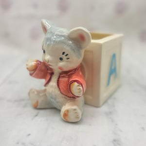 May include: A vintage ceramic figurine of a bear cub wearing a red jacket, seated next to a cream-colored block with the letter "A" in blue. The bear has blue and white fur and is holding its paw up.