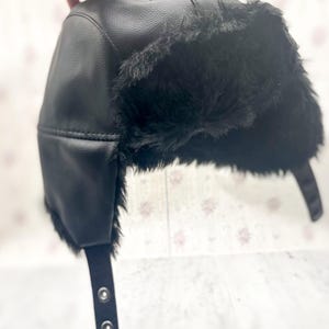 May include: Black faux leather trapper hat with black faux fur trim. The hat features ear flaps and a chin strap with snap closures. A stylish and warm winter accessory.
