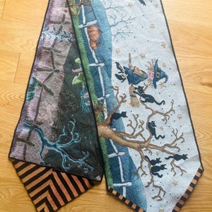 May include: A decorative Halloween-themed table runner. The runner features a tapestry design with witches, black cats, and bats. The color palette includes black, orange, blue, and white. The runner has a black border and tassels at the ends.