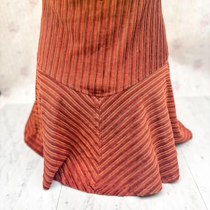 May include: A corduroy skirt in shades of red and brown. The skirt features vertical stripes at the waist and a chevron pattern on the lower half. The fabric has a soft, textured appearance, and the skirt has a flared silhouette.