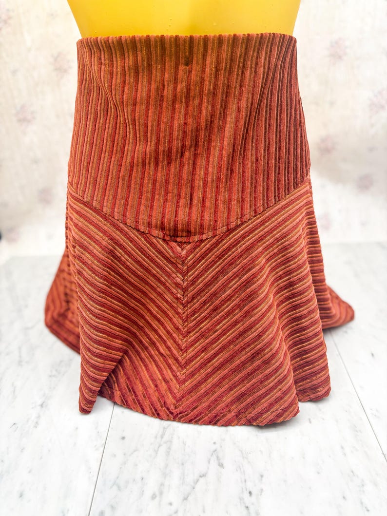 May include: A corduroy skirt in shades of red and brown. The skirt features vertical stripes at the waist and a chevron pattern on the lower half. The fabric has a soft, textured appearance, and the skirt has a flared silhouette.