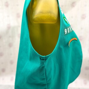 May include: A teal vest with a scoop neck and a rounded hem. The vest has a patch with the number "901" and a green emblem. An orange and yellow curved patch is also visible. The vest is displayed on a mannequin.