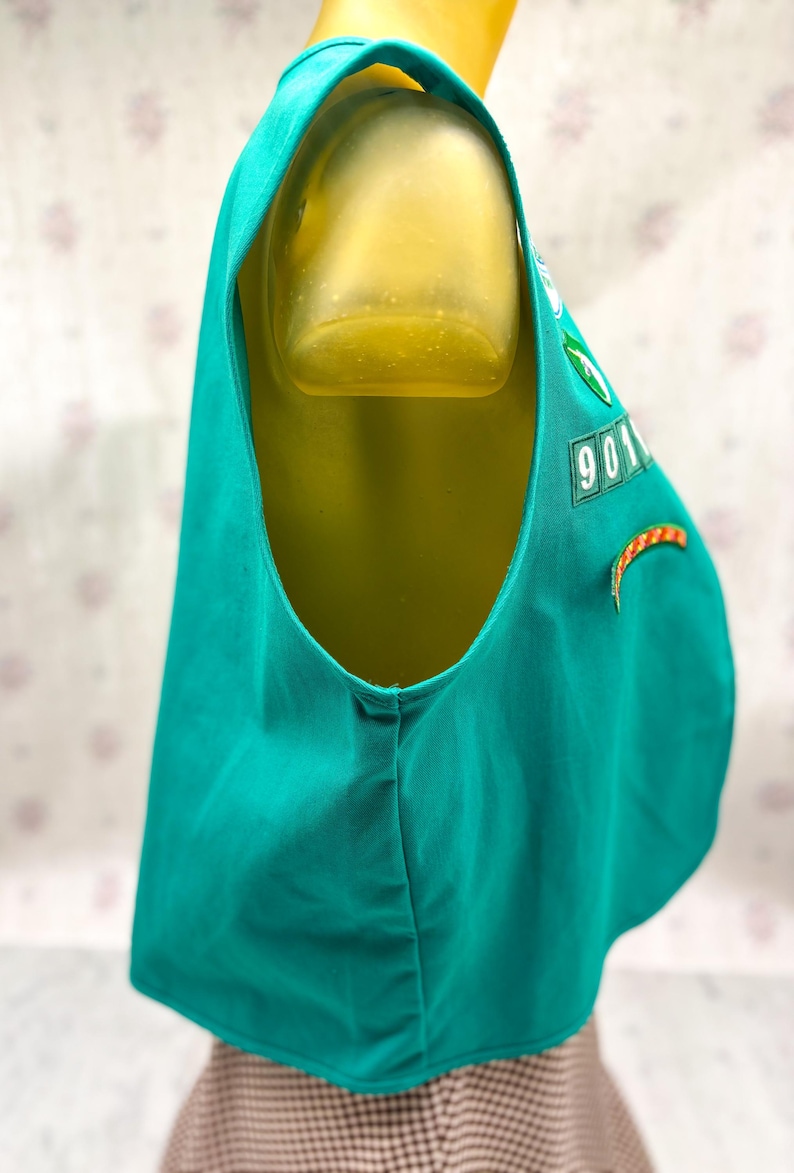 May include: A teal vest with a scoop neck and a rounded hem. The vest has a patch with the number "901" and a green emblem. An orange and yellow curved patch is also visible. The vest is displayed on a mannequin.