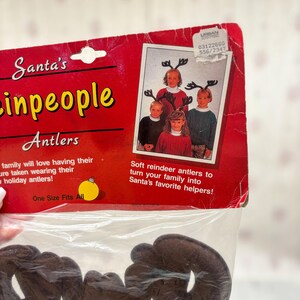 May include: A package of brown reindeer antlers for holiday celebrations. The packaging is red with the text "Santa's inpeople Antlers" and a photo of children wearing the antlers. The text "One Size Fits All" is also visible.