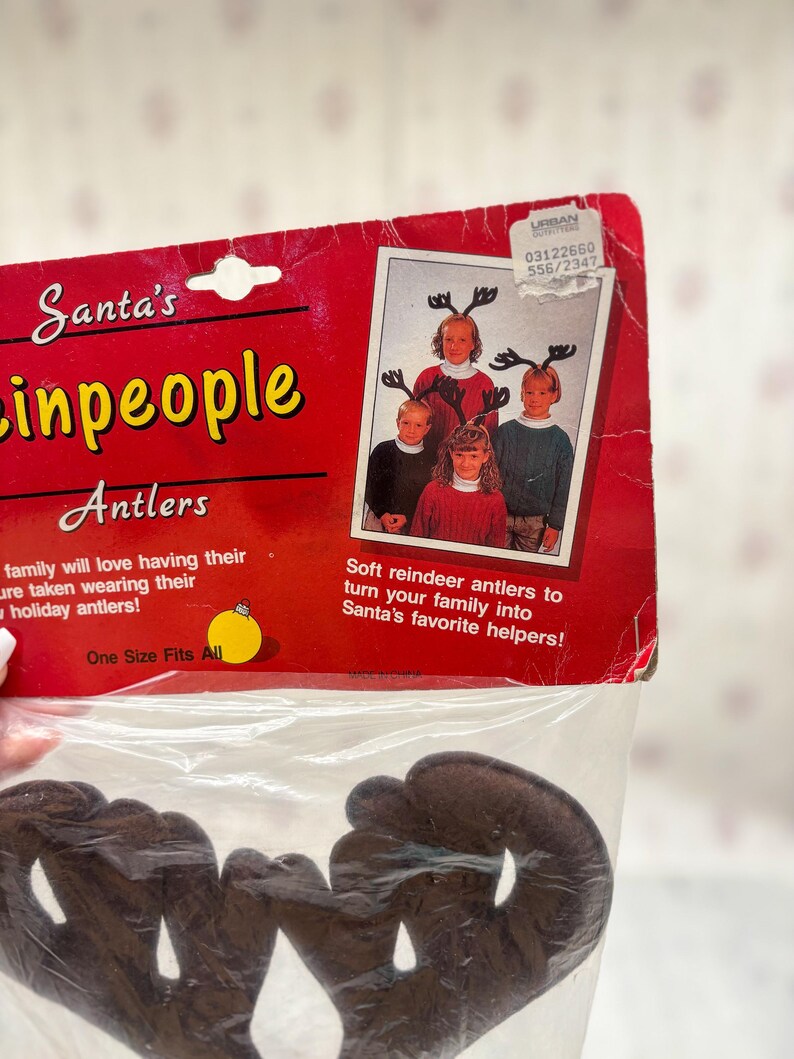 May include: A package of brown reindeer antlers for holiday celebrations. The packaging is red with the text "Santa's inpeople Antlers" and a photo of children wearing the antlers. The text "One Size Fits All" is also visible.