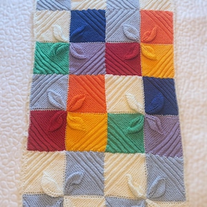 May include: A knitted blanket featuring a patchwork design with squares in various colors, including blue, green, orange, and yellow. Each square has a leaf-shaped embellishment. The blanket is made of soft yarn and is suitable for a baby.