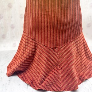 May include: A corduroy skirt in shades of red and brown. The skirt features a high waist and a flared, asymmetrical hemline. The fabric has a ribbed texture, with vertical stripes at the waist and diagonal stripes at the hem.