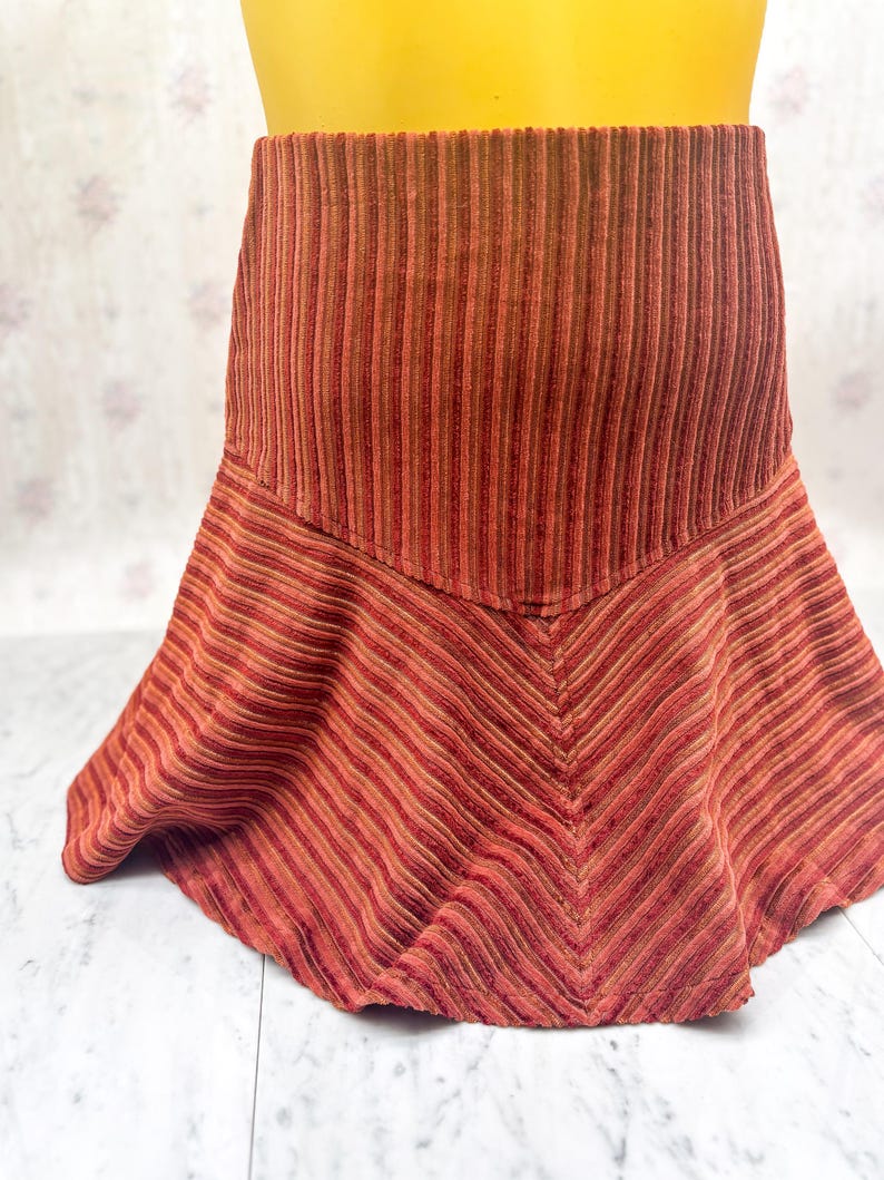 May include: A corduroy skirt in shades of red and brown. The skirt features a high waist and a flared, asymmetrical hemline. The fabric has a ribbed texture, with vertical stripes at the waist and diagonal stripes at the hem.