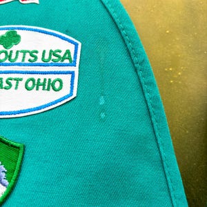 May include: Close-up of a teal green fabric with embroidered patches. Patches include a US flag, a "SCOUTS USA" patch, and a "EAST OHIO" patch. The fabric has a stitched seam along the right side.
