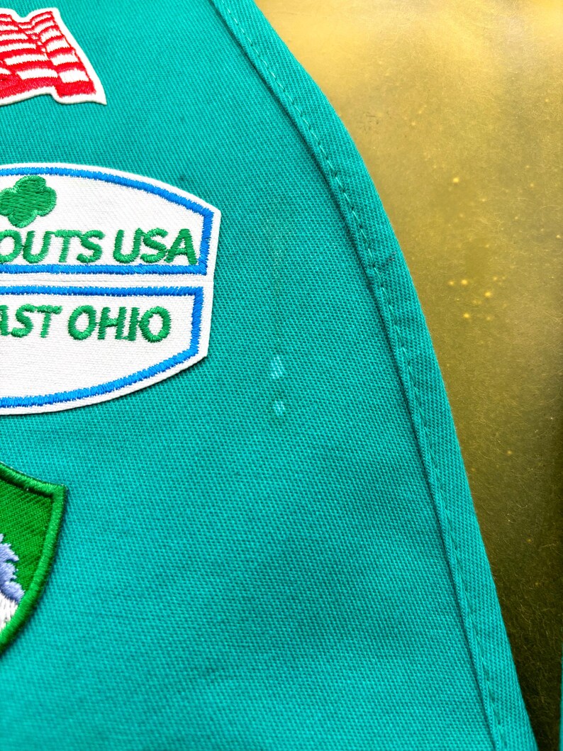 May include: Close-up of a teal green fabric with embroidered patches. Patches include a US flag, a "SCOUTS USA" patch, and a "EAST OHIO" patch. The fabric has a stitched seam along the right side.