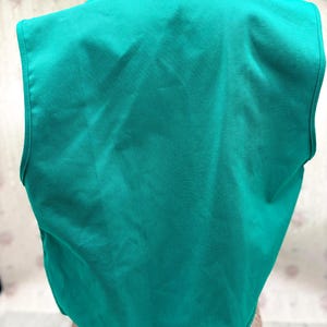 May include: A teal-colored vest with a V-neck design. The vest is sleeveless and appears to be made of a smooth fabric. The back of the vest is visible, showing the full shape and cut of the garment. The vest is a solid color.