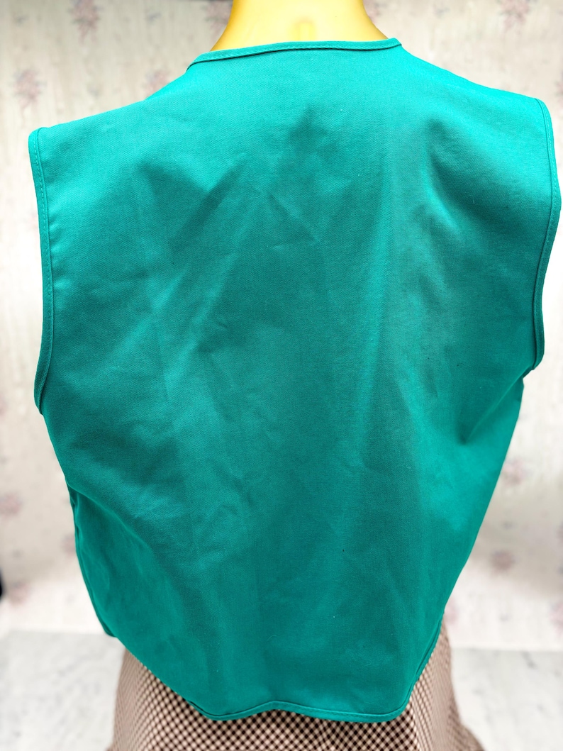 May include: A teal-colored vest with a V-neck design. The vest is sleeveless and appears to be made of a smooth fabric. The back of the vest is visible, showing the full shape and cut of the garment. The vest is a solid color.
