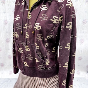 May include: A maroon zip-up hoodie with a hood and front pockets. The hoodie is patterned with a repeating gold-colored "SP" logo. The zipper and drawstring tips are gold. The hoodie is displayed on a mannequin.
