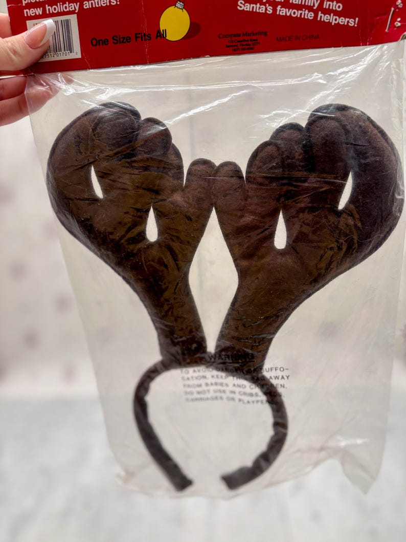 May include: Brown reindeer antler headband in a clear plastic package. The antlers are made of a soft, plush material and are attached to a dark brown headband. The packaging reads "One Size Fits All".