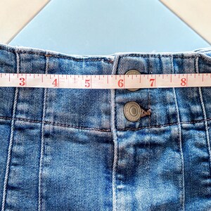 May include: Close-up of a light blue denim skirt with a measuring tape across the waistband. The skirt has a button closure and vertical seam details. The waistband measures approximately 12 inches.