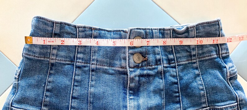 May include: Close-up of a light blue denim skirt with a measuring tape across the waistband. The skirt has a button closure and vertical seam details. The waistband measures approximately 12 inches.