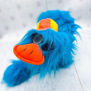 May include: A plush toy featuring long, bright blue faux fur, a prominent orange beak, and a single large, round eye. The creature wears a small, rounded cap with alternating yellow and orange panels positioned on the top of its head.