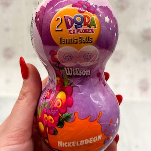 May include: A purple and orange container of Wilson tennis balls featuring the Nickelodeon Dora the Explorer logo. The container has a unique hourglass shape and is decorated with floral and star designs.