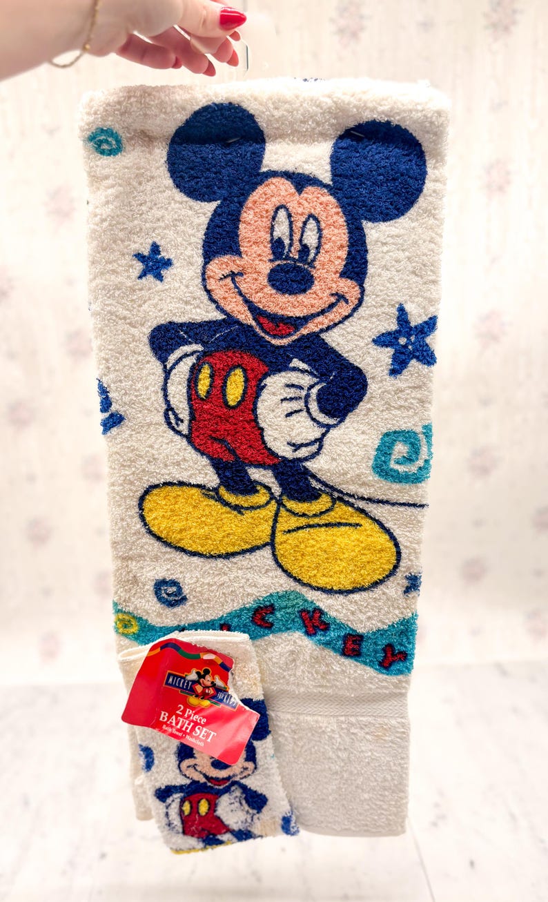 May include: White Mickey Mouse 2-piece bath set. The towel features a large image of Mickey Mouse in his classic outfit, with blue stars and a blue and turquoise border. The set includes a matching washcloth.