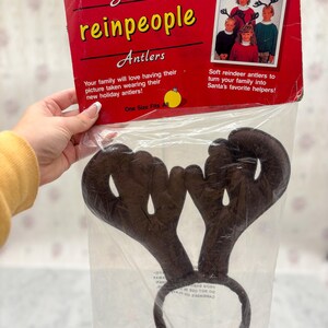 May include: Brown reindeer antler headband in a clear plastic package. The package is red and has the text "Santa's reinpeople Antlers". The text on the package says "One Size Fits All".