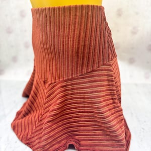 May include: A rust-colored, corduroy skirt with vertical stripes. The skirt has a high waist and a flared, asymmetrical hemline. The fabric appears to have a slight sheen, and the color is a mix of red, orange, and brown tones. The skirt is displayed on a mannequin.