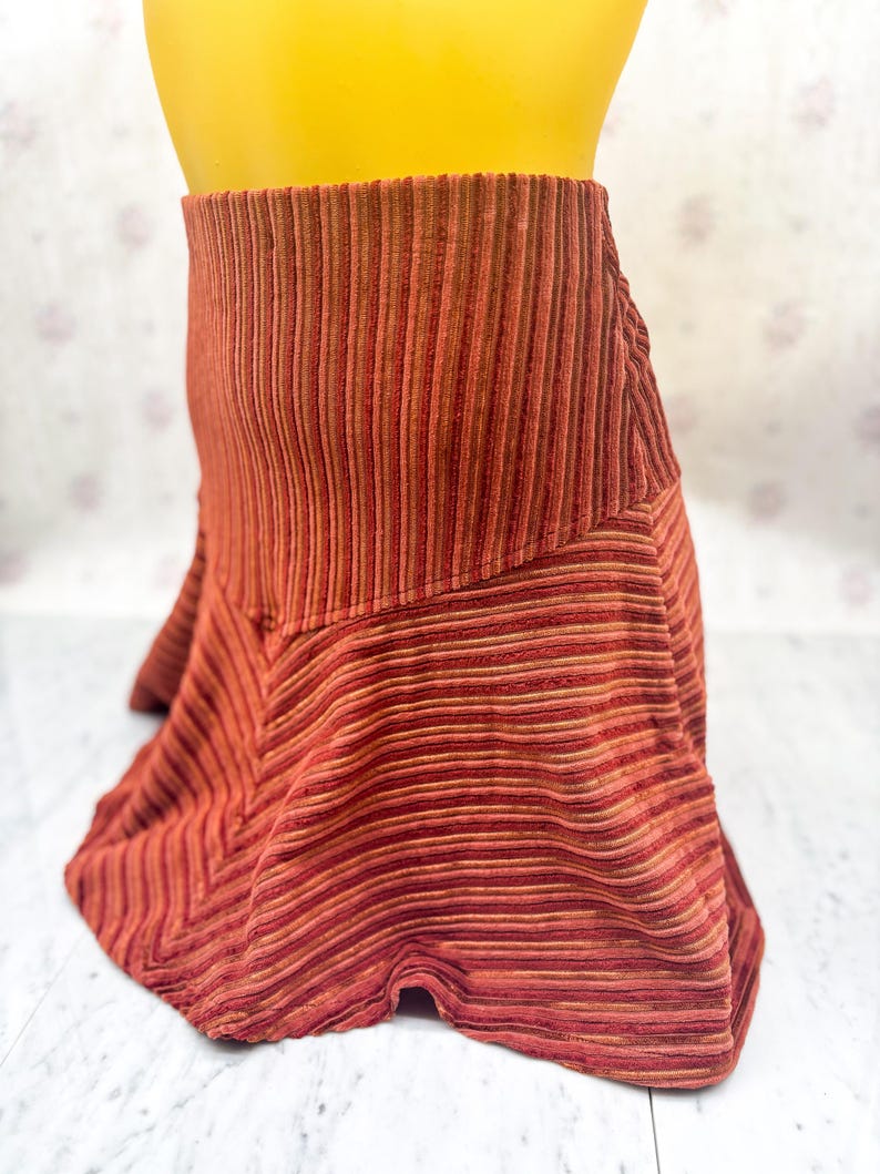 May include: A rust-colored, corduroy skirt with vertical stripes. The skirt has a high waist and a flared, asymmetrical hemline. The fabric appears to have a slight sheen, and the color is a mix of red, orange, and brown tones. The skirt is displayed on a mannequin.