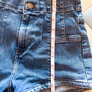 May include: Close-up of blue denim shorts with a high waist and two button closure. The shorts are measured at approximately 12 inches in length. The shorts have a classic five-pocket design.