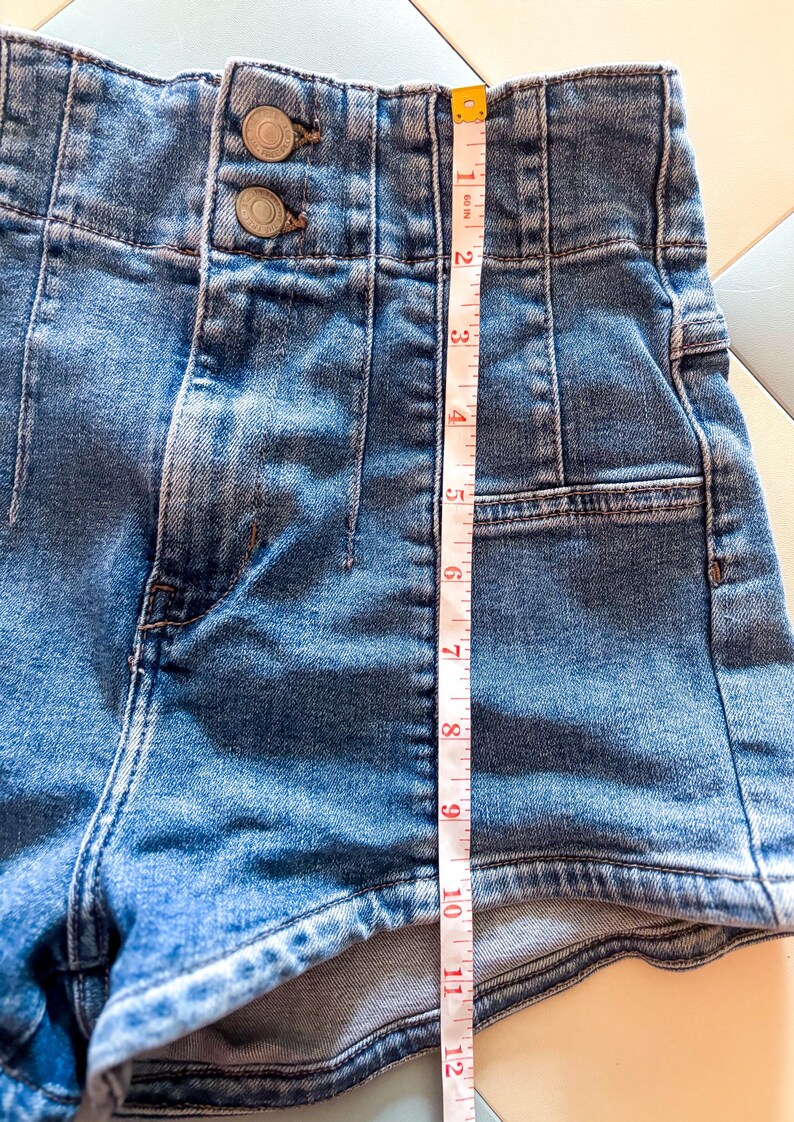 May include: Close-up of blue denim shorts with a high waist and two button closure. The shorts are measured at approximately 12 inches in length. The shorts have a classic five-pocket design.