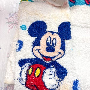 May include: White terry cloth towel featuring a smiling Mickey Mouse design. Mickey is depicted with blue ears and head, a red body, and yellow gloves. The towel has a textured, plush appearance.