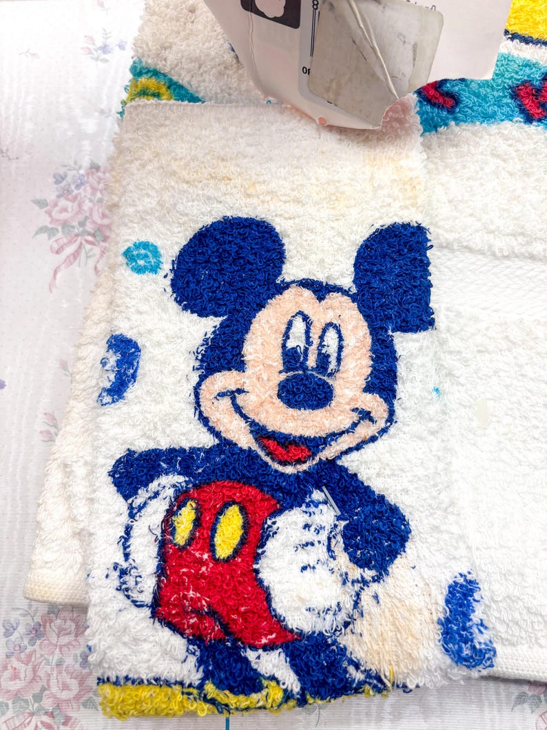 May include: White terry cloth towel featuring a smiling Mickey Mouse design. Mickey is depicted with blue ears and head, a red body, and yellow gloves. The towel has a textured, plush appearance.