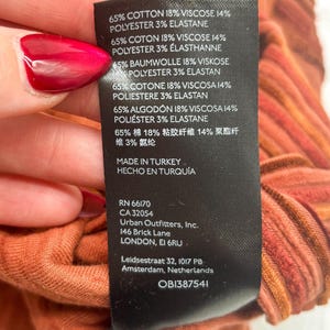 May include: Close-up of a clothing tag with fabric composition details: 65% cotton, 18% viscose, 14% polyester, and 3% elastane. The tag also includes the manufacturer's information and the text "MADE IN TURKEY". The fabric is a reddish-orange color.