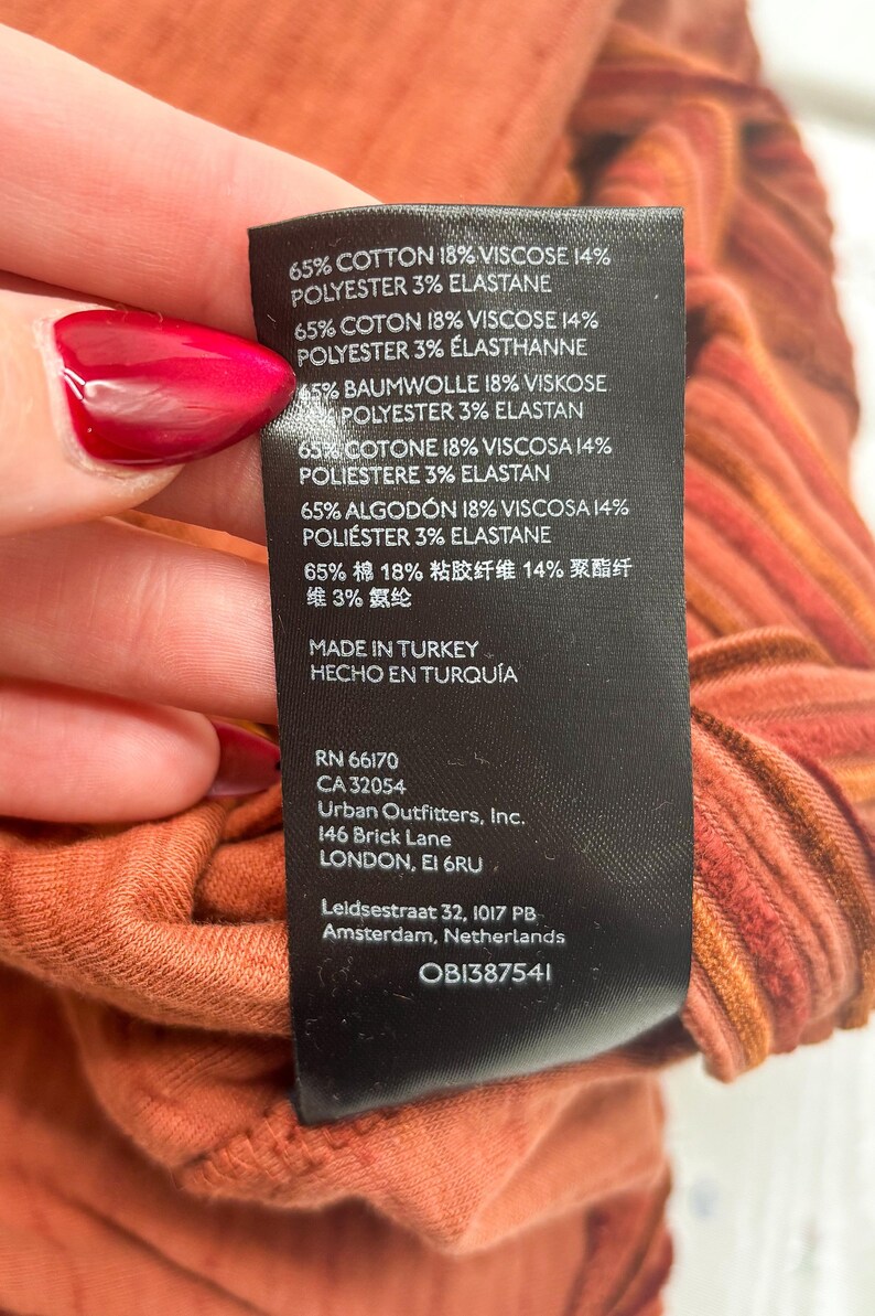 May include: Close-up of a clothing tag with fabric composition details: 65% cotton, 18% viscose, 14% polyester, and 3% elastane. The tag also includes the manufacturer's information and the text "MADE IN TURKEY". The fabric is a reddish-orange color.