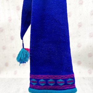 May include: A vibrant blue scarf with a decorative pattern in pink and teal near the bottom edge. The scarf has a tassel with teal and pink accents. The scarf is made of a woven material.