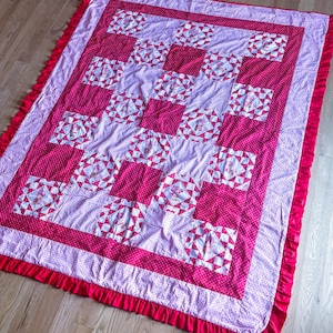 May include: A handmade quilt in pink and white, with a patchwork design. The quilt has a repeating pattern of squares and triangles, and a red ruffle border. A cosy and decorative item for a bed or sofa, with a traditional feel.