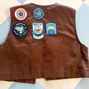 May include: A brown vest with several patches. Patches include text such as "Native America", "Incredible Voyage Cookies '95", "Friendships Around The World '95", "MMGSC '94 Partners For Tomorrow Calendar-Nut Sale", and "Booth Sales 1995".