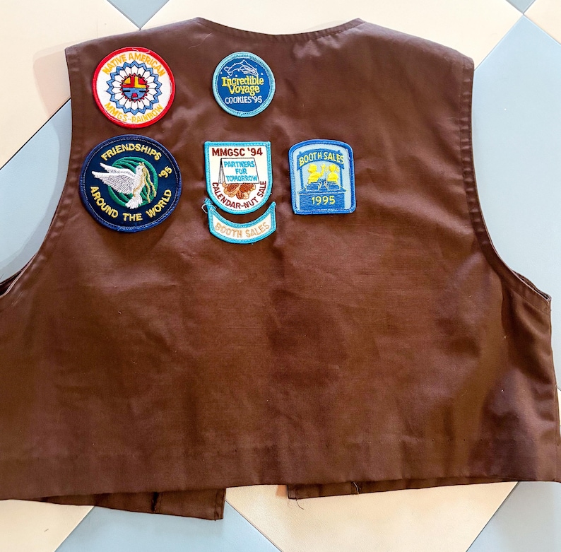 May include: A brown vest with several patches. Patches include text such as "Native America", "Incredible Voyage Cookies '95", "Friendships Around The World '95", "MMGSC '94 Partners For Tomorrow Calendar-Nut Sale", and "Booth Sales 1995".